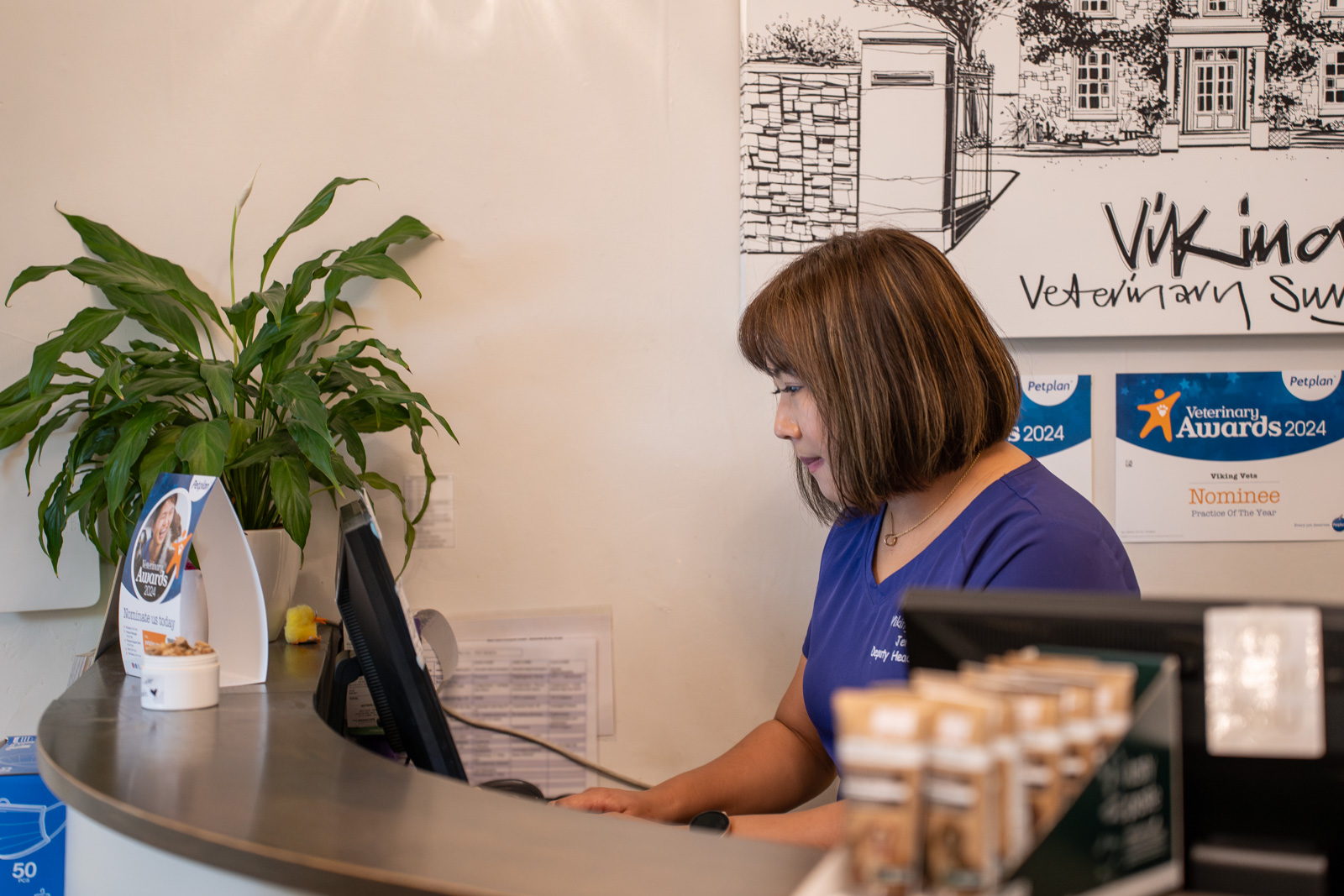 A woman in a purple shirt works at a computer behind a reception desk in a veterinary clinic. Nearby are brochures and a plant. Awards for 2024 are displayed on the wall in the background.