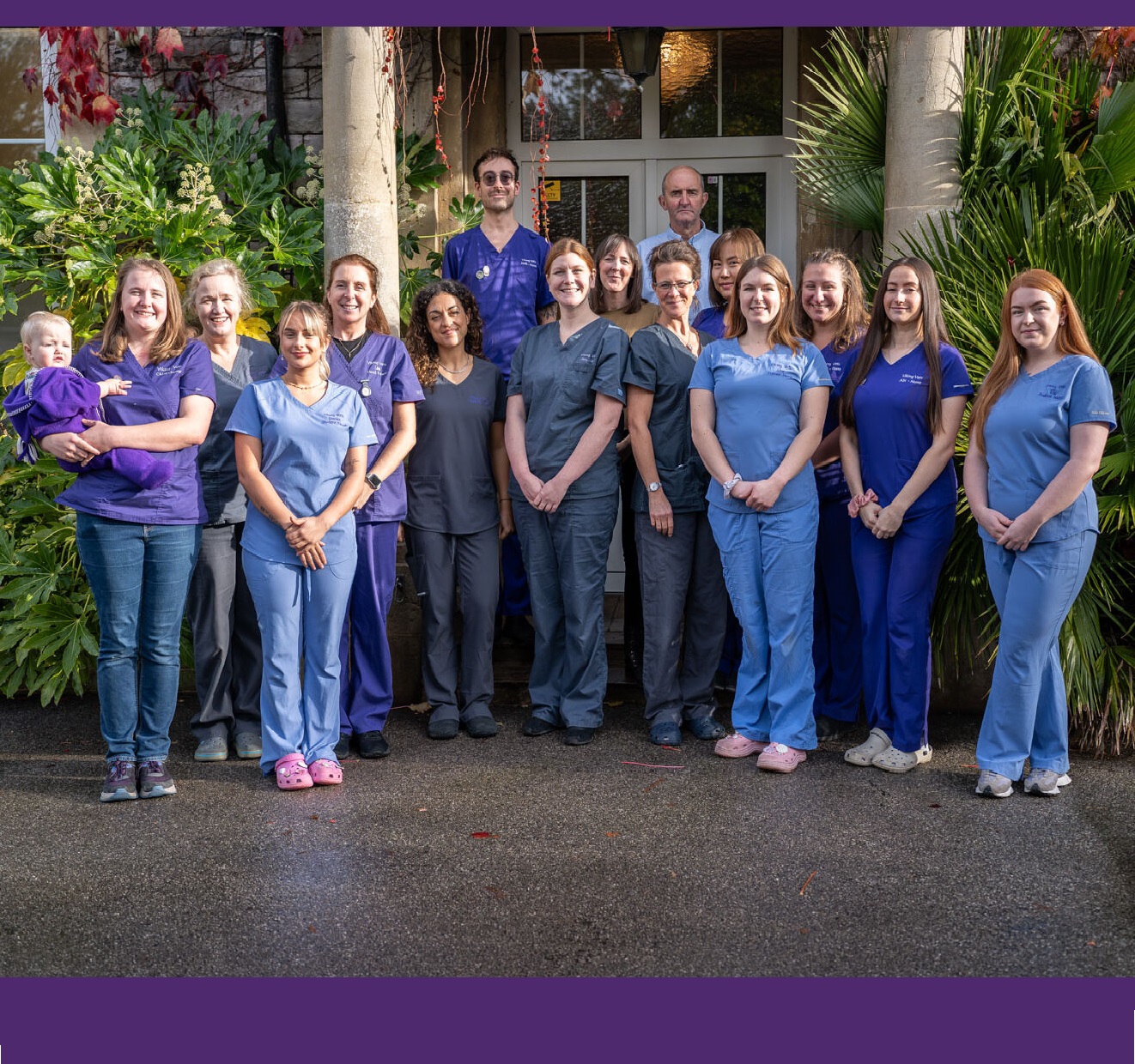 A group of sixteen people, some in blue and purple scrubs, standing and smiling in front of a building entrance surrounded by greenery. One person on the left is holding a baby.