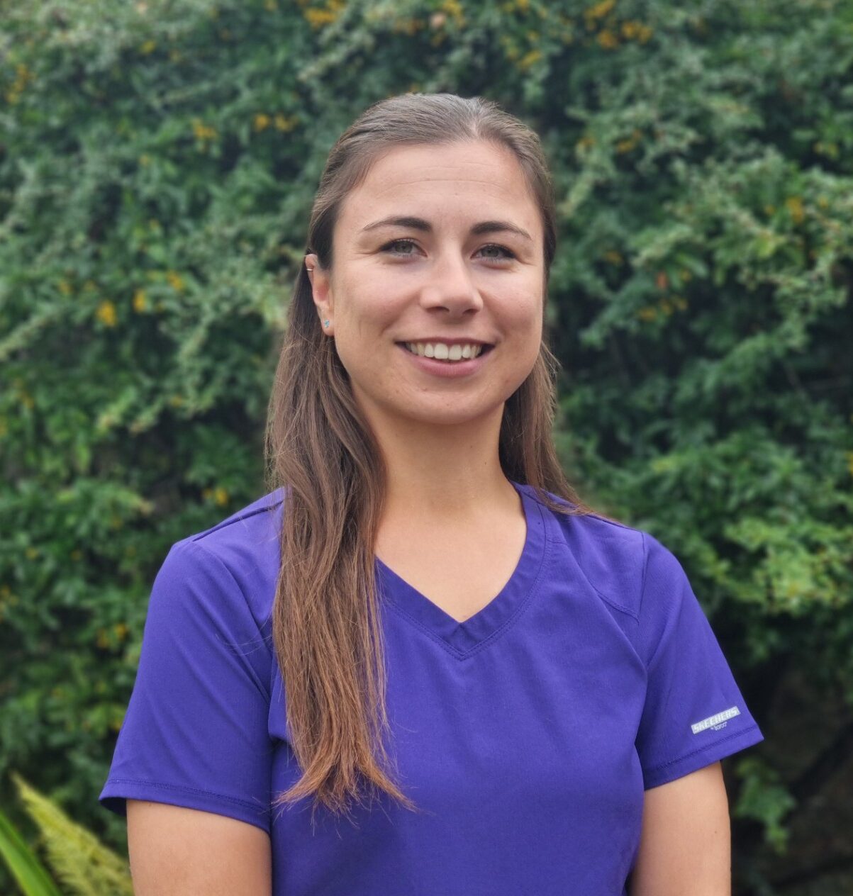 A woman with long brown hair wearing a purple scrub top is standing outdoors, smiling, with green foliage in the background.